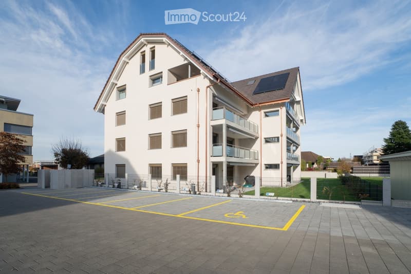 Modern multi-story residential building with white facade, multiple windows, balconies, and solar panels on the roof. The building is surrounded by a paved parking area with designated spaces.