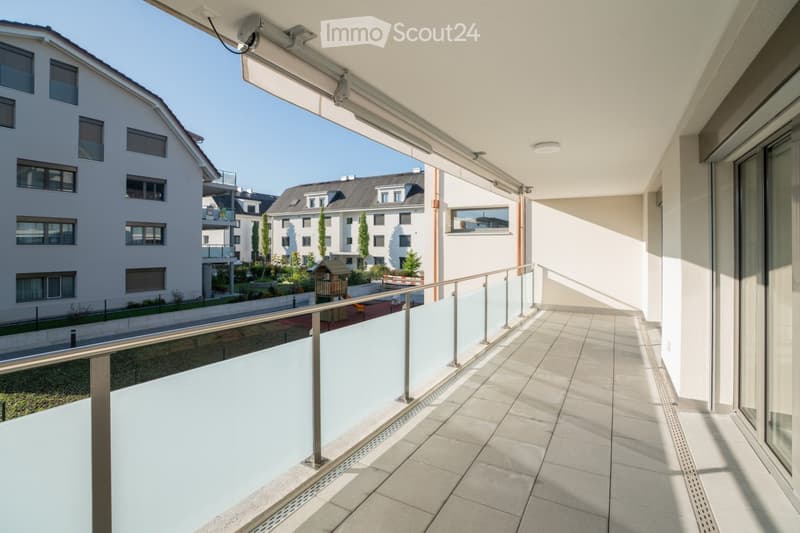 Modern balcony with tiled floor, metal railing, and retractable awning, overlooking a residential area with buildings and greenery.