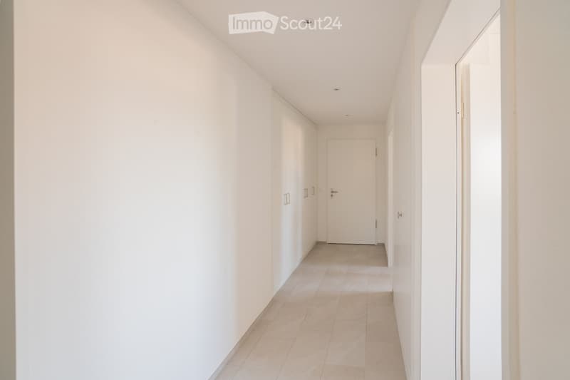A clean, modern hallway with white walls, tiled flooring, and recessed ceiling lighting. Features a closed door at the end and built-in storage units along one wall.
