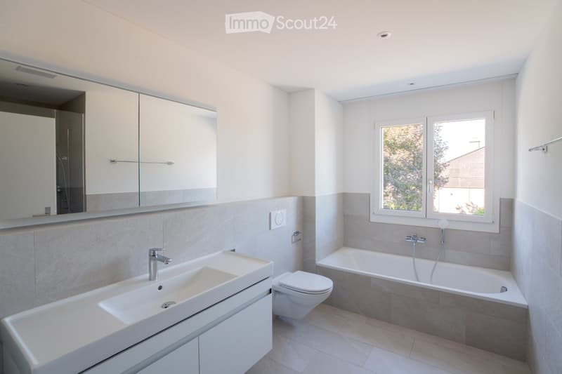 Modern bathroom with a large sink, toilet, bathtub, and mirrored cabinet. Light-colored tiles and ample natural light from a window.