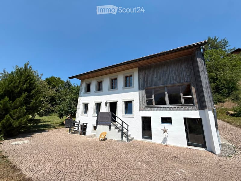 Two-story house with white walls, wooden accents, multiple windows, and a raised ground floor entrance with steps.