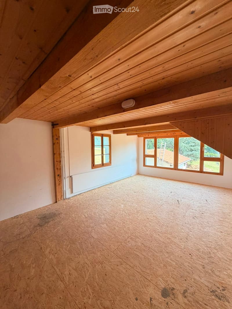 Empty attic space with wooden beams, exposed wooden ceiling, wooden floor, and windows letting in natural light.