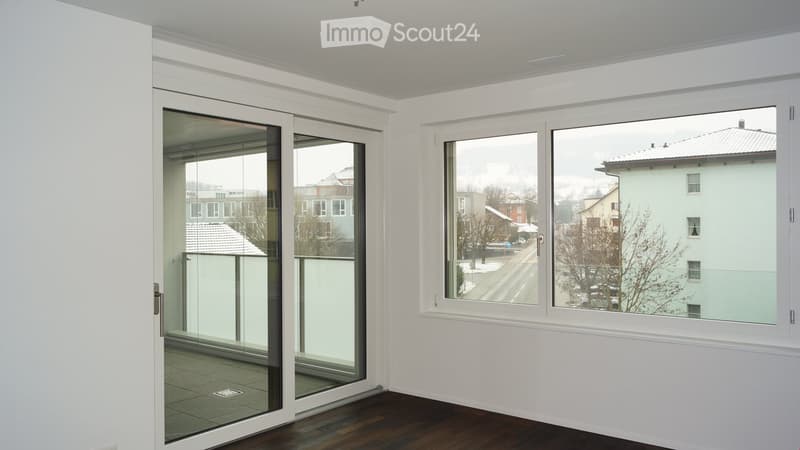 Empty room with white walls, wooden floor, large windows, and a sliding glass door leading to a balcony.