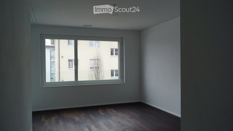 Empty room with wooden flooring, white walls, and a large window offering a view of neighboring buildings.