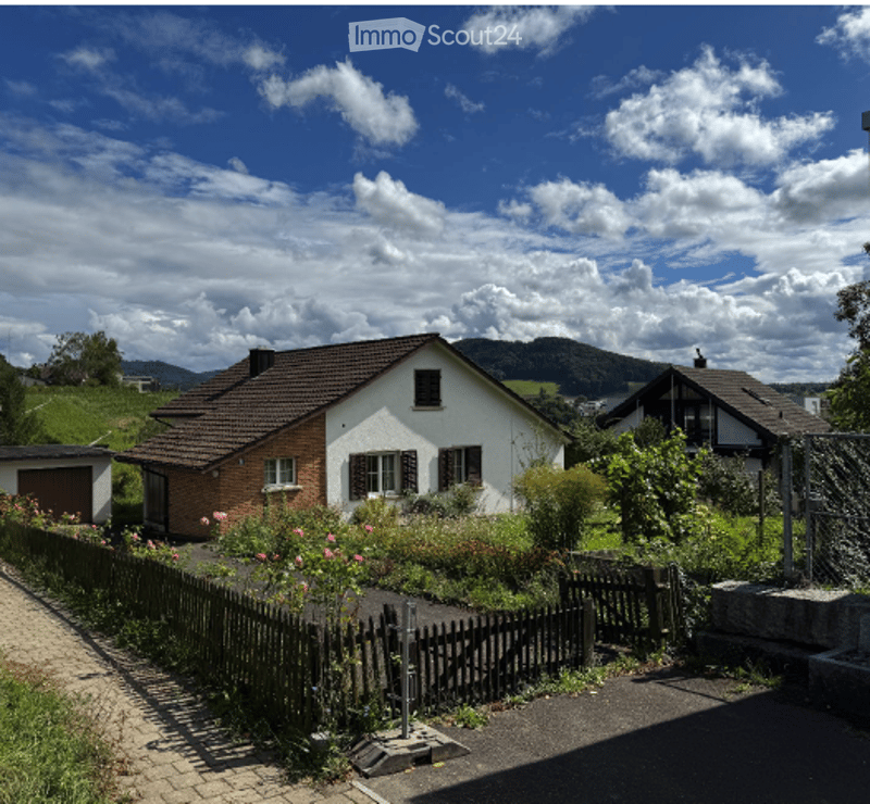 A two-story house with a brown tiled roof and white walls, surrounded by a garden with colorful flowers and a wooden fence. The house features multiple windows and a small garage attached to the left side.