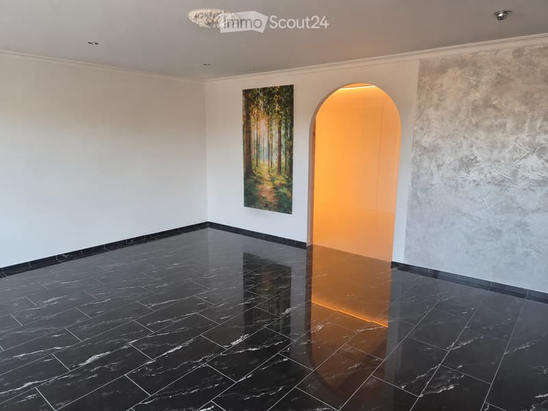Empty hallway with black marble flooring, white walls, and a textured wall on the right. An arched doorway leads to a warmly lit area, and a painting of a forest scene hangs on the left wall.