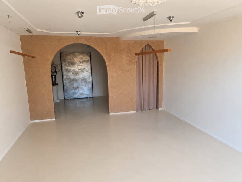 Empty room with beige walls, polished concrete floor, and a large arched doorway leading to another space. Minimalist decor with a single abstract artwork and a small decorative item visible in the background.