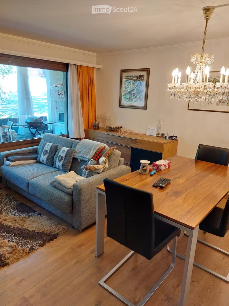 Living room with a grey sofa, wooden dining table, chairs, chandelier, and large window with a view.