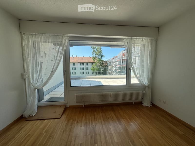 Empty room with wooden flooring, large window with white curtains, radiator, and a view of a balcony and cityscape.