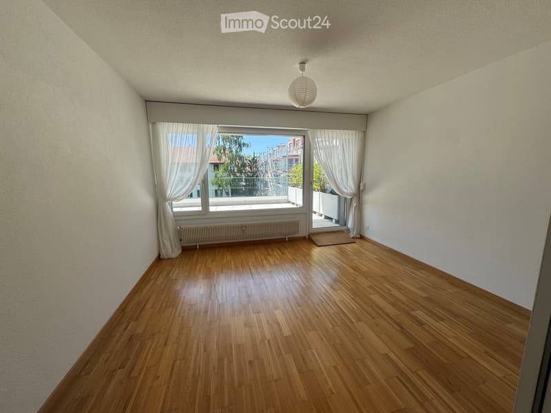 Empty room with wooden flooring, white walls, large window with white curtains, radiator, and a hanging light fixture.