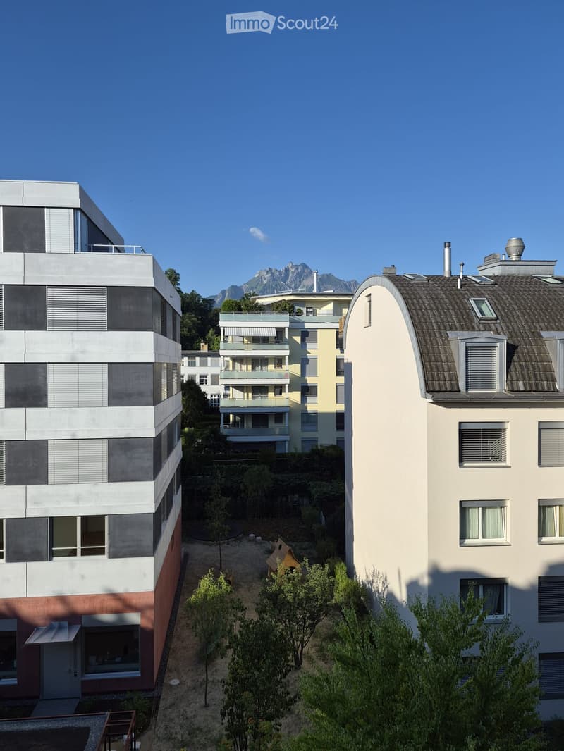 Modern multi-story residential buildings with balconies and large windows, surrounded by greenery and a clear blue sky with mountain views.