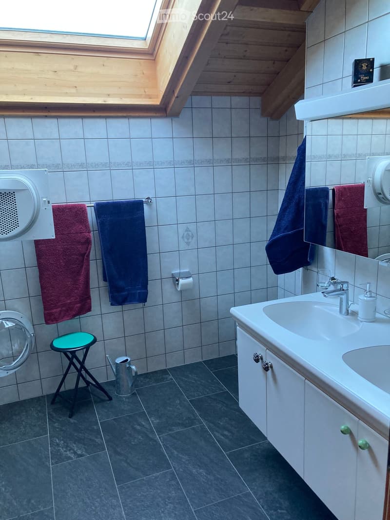 Bathroom with a white sink, mirror, towel rack with towels, a small green stool, and a toilet paper holder. The walls are tiled, and there is a skylight window at the top.