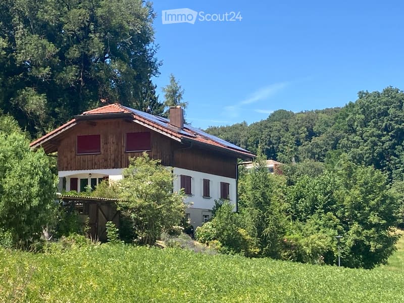 A two-story wooden house with a red-tiled roof, surrounded by lush greenery and trees.