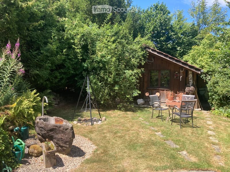 A serene garden area with a wooden cabin, outdoor seating, a fire pit, and lush greenery.