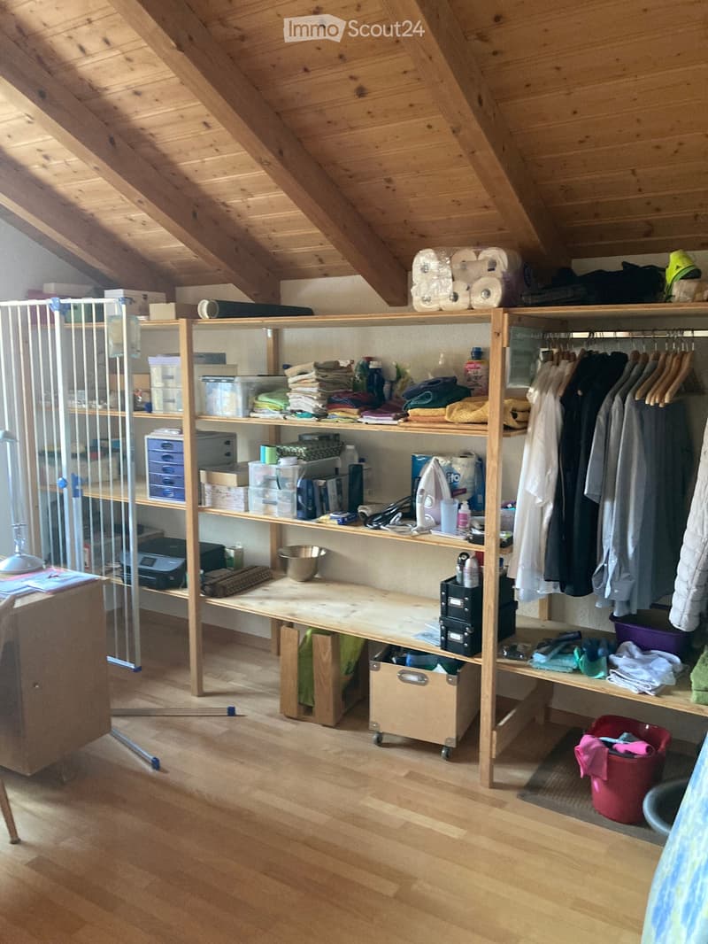 Attic storage room with wooden slanted ceilings, wooden shelves filled with folded clothes, cleaning supplies, and storage boxes. A clothing rack with hanging garments is on the right, and a desk with a lamp is on the left.