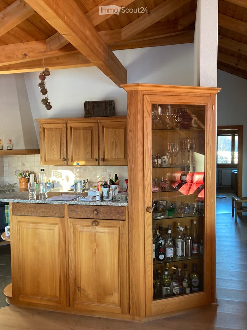 A wooden kitchen with a built-in bar area, featuring cabinets, a countertop, and a display cabinet filled with bottles and glasses.