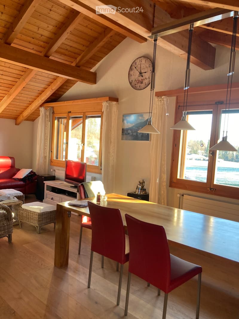 Bright, airy room with wooden beams, large windows, a red leather sofa, a wooden dining table with red chairs, a television, and a decorative clock on the wall.