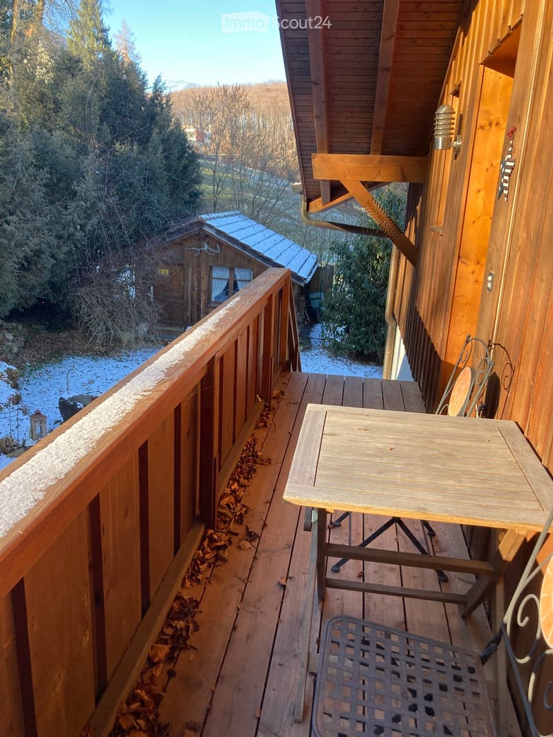 Wooden balcony with a table and chair, overlooking a snowy landscape with trees and a small wooden structure.