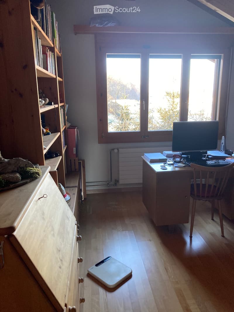 Bright room with wooden floor, large windows, desk with computer, bookshelf, and a small stool on the floor.