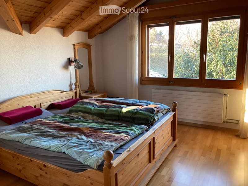 Cozy bedroom with wooden bed, colorful quilt, wooden nightstand, mirror, and wall-mounted lamp. Large windows with wooden frames and natural light.