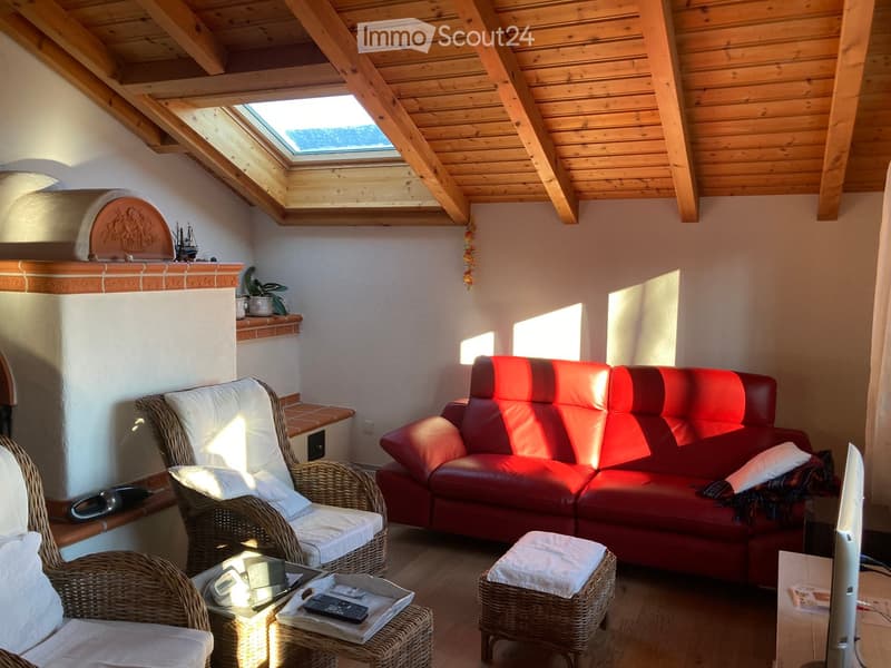 Cozy living room with a red leather sofa, wicker chairs, a small table, and a skylight window. The room features a fireplace with a decorative mantel and a wooden ceiling.