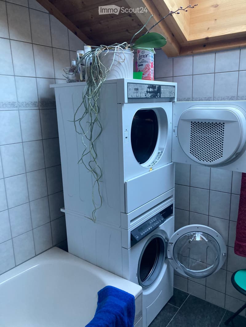 Bathroom with a washing machine and dryer stacked, a small stool, and a plant on top of the washing machine.