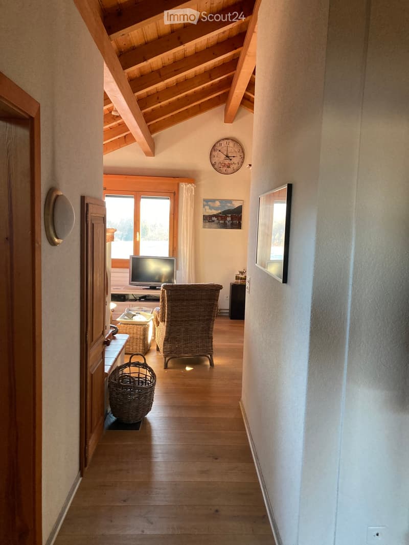 A wooden-floored hallway with exposed wooden beams on the ceiling, leading to a living area with a wicker chair, TV, and small table.