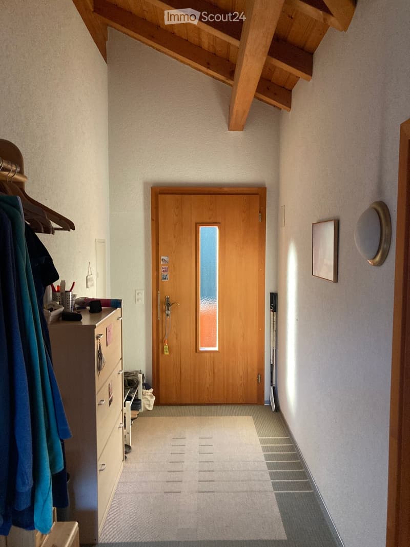 A narrow hallway with a wooden door at the end, a coat rack with clothes, a small table with various items, and a mirror on the wall.