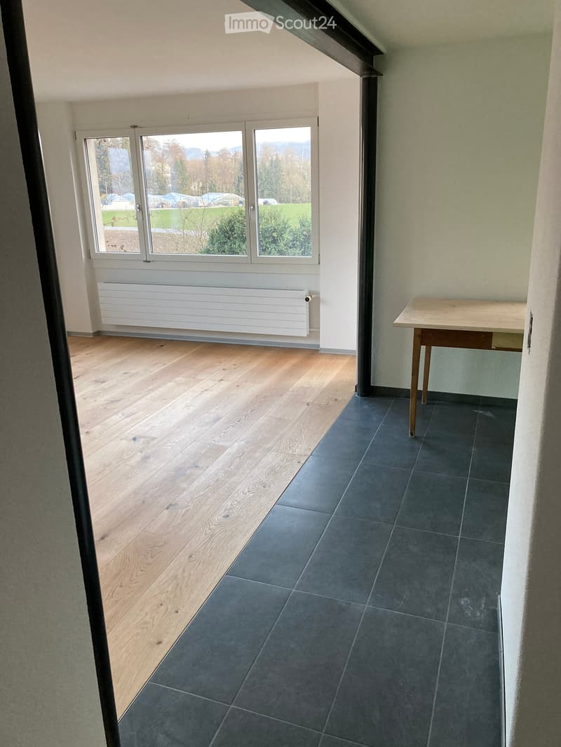 A small, unfurnished room with wooden flooring, a radiator, and a table near the window. The room has large windows offering a view of the outdoors.
