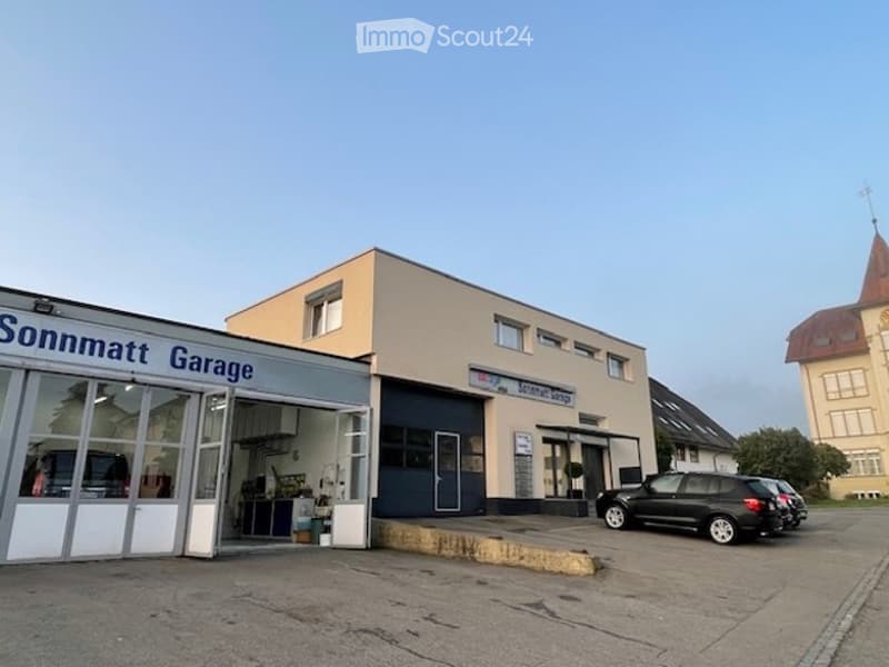 A modern, single-story building with a garage entrance labeled "Sonnmatt Garage." The building has a flat roof, large windows, and a clean, contemporary design. A black SUV is parked in front of the garage.