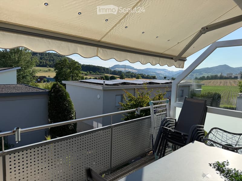 Balcony with a large retractable awning, metal railing, and outdoor furniture including a table and chairs, overlooking a scenic countryside view.