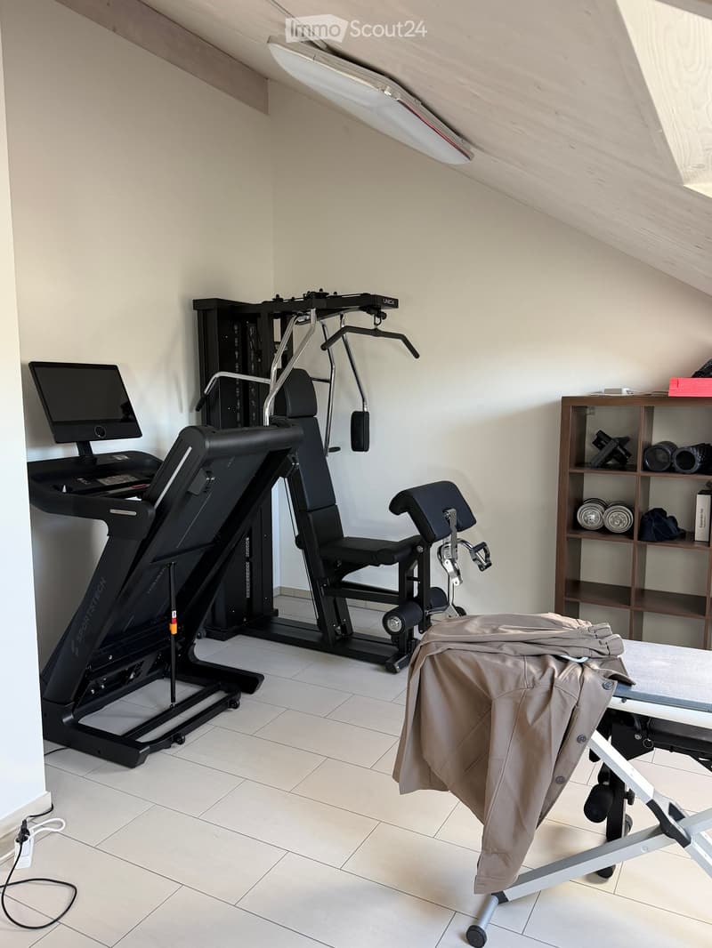 A well-equipped home gym with a treadmill, weight machine, chair, table, shelf, laptop, and camera. The room has white walls and a slanted ceiling typical of an attic space.
