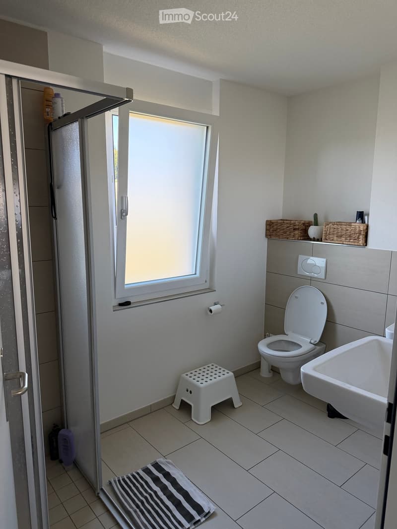 Modern bathroom with white tiled walls and floor, featuring a toilet, bathtub, small white stool, and a striped towel on the floor. A wicker basket with a small plant sits on a shelf above the toilet.