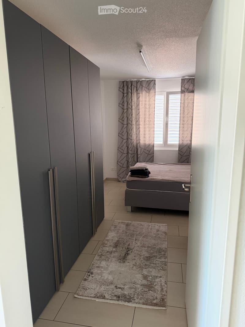 Modern bedroom with a bed, a large rug, and patterned curtains. The room features tiled flooring and built-in dark-colored wardrobes.