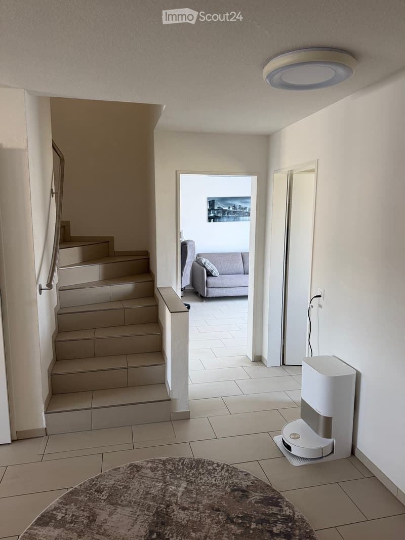 Hallway with a round table, a robot vacuum, and a staircase leading to a living room with a sofa and a cityscape painting.