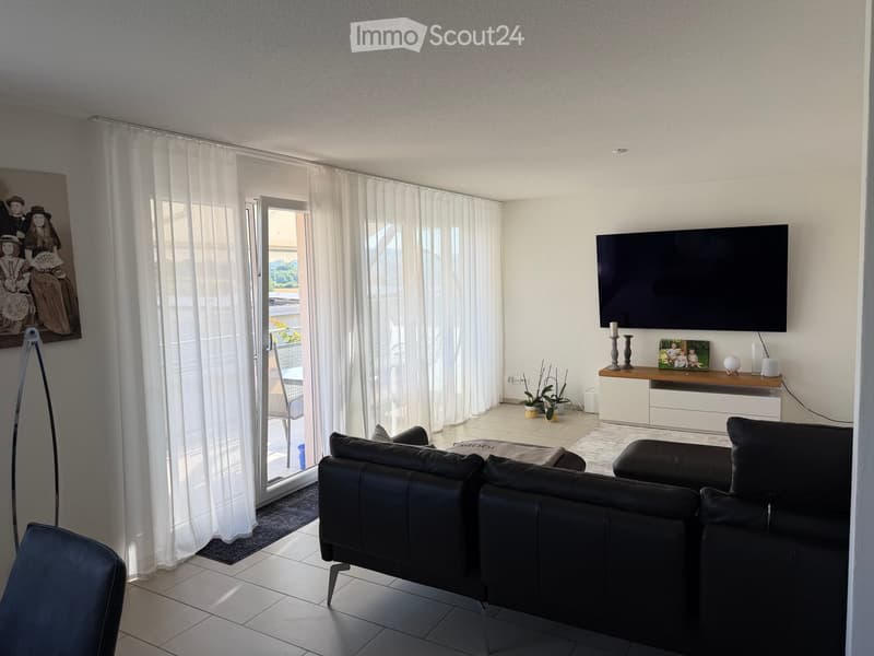 Modern living room with large windows, white curtains, black leather sofa, ottoman, chair, TV, and decorative items on a console table.