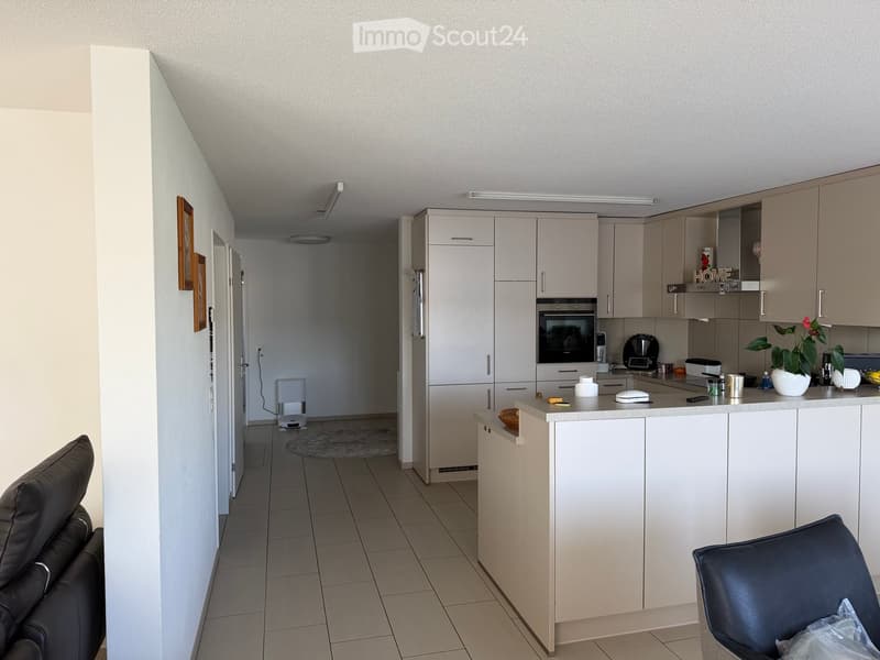 Modern kitchen with light-colored cabinets, a central island, and a living area with a sofa and chair. The kitchen features an oven, coffee maker, and a small plant on the counter.