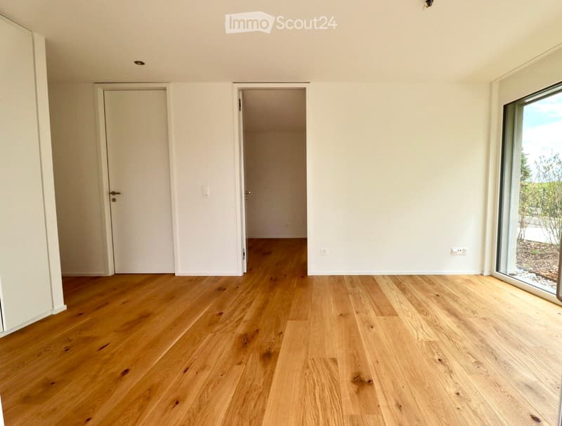Empty room with light wooden flooring, white walls, and large windows offering a view of the outdoors.