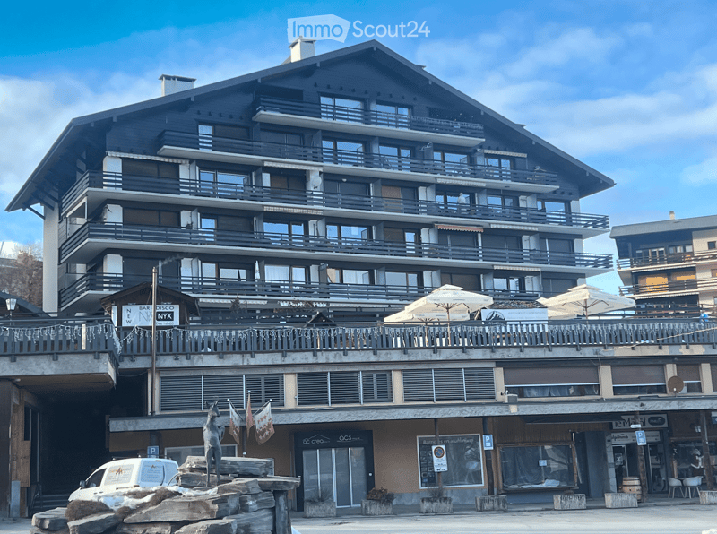 Multi-story building with balconies, large windows, and a restaurant area on the ground floor. Features include outdoor seating, flags, and a decorative statue.