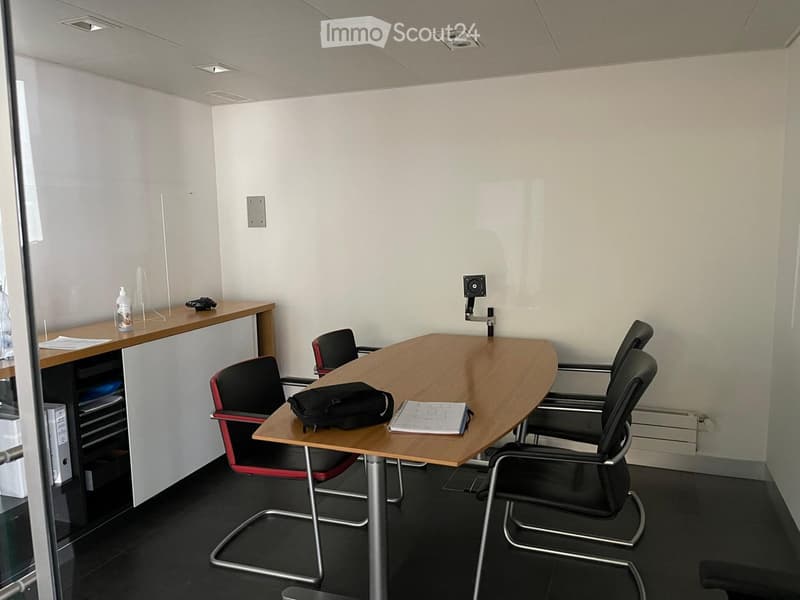 A modern office space with a large wooden table, several black chairs, a backpack, a bottle, and a screen mounted on the wall.