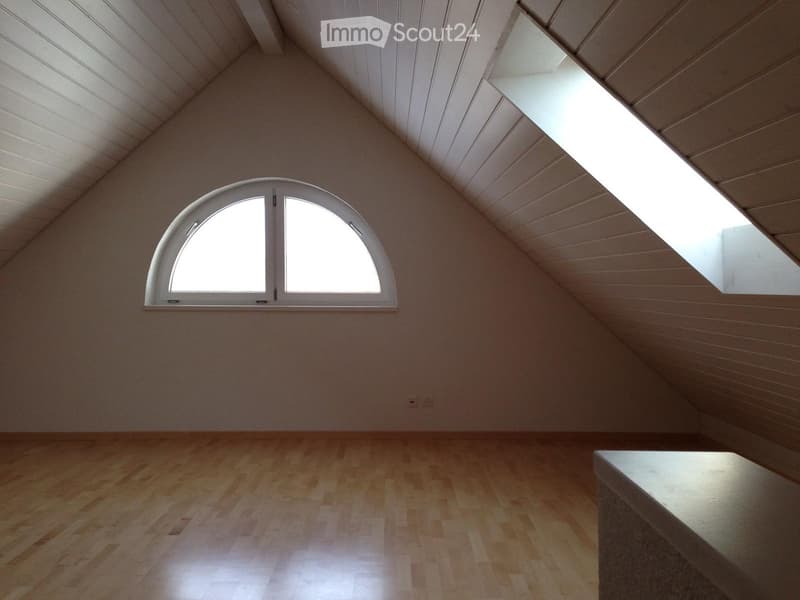 Empty attic room with wooden flooring, slanted wooden ceiling, and large arched window allowing natural light.