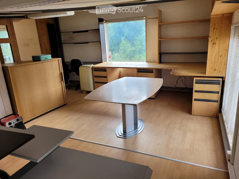 A modern, well-lit office space with wooden flooring, large windows, a central round table, chairs, cabinets, and shelves.