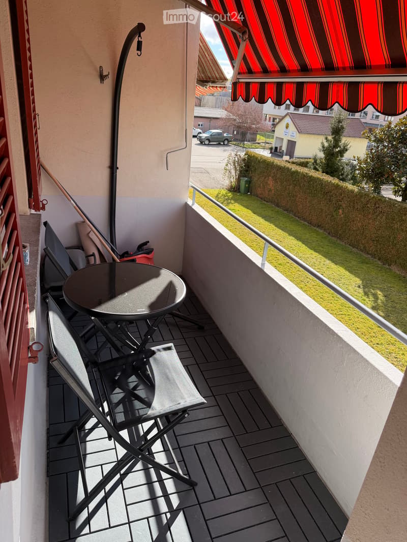 Balcony with tiled floor, table, and chairs, featuring a view of a grassy area and nearby buildings.