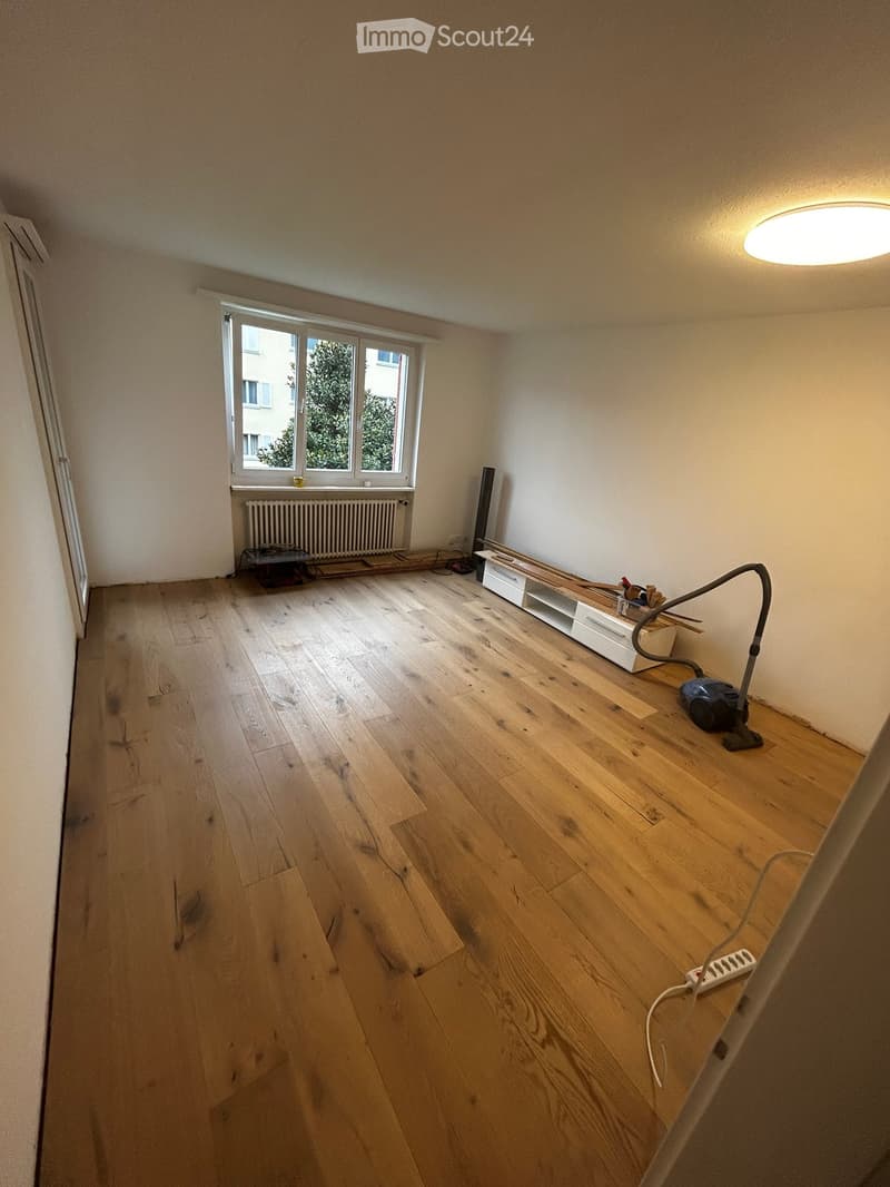 Empty room with wooden flooring, white walls, and a large window with a radiator below it. The room is clean and unfurnished, with only a few scattered items like a vacuum cleaner and some tools.