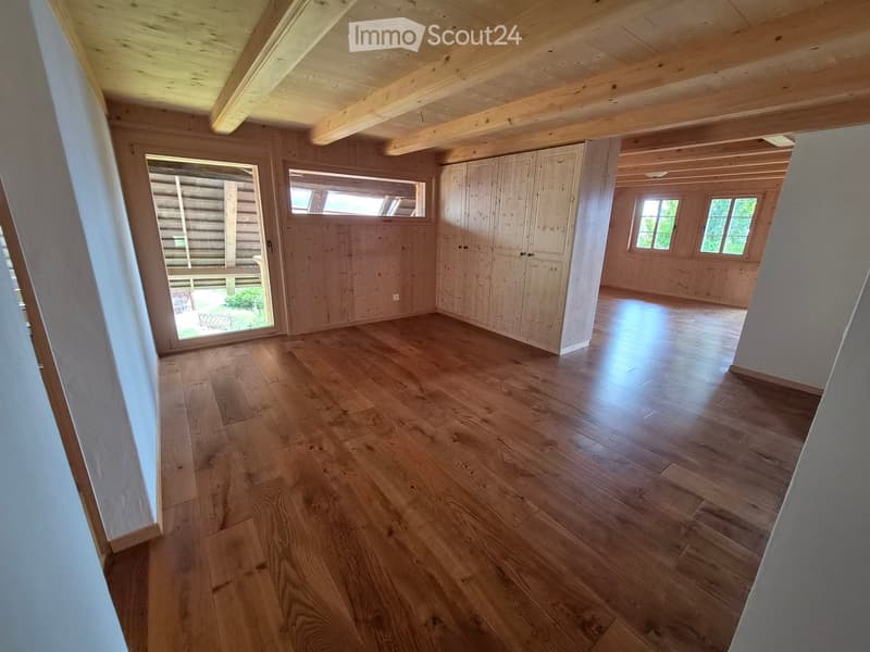 Empty room with wooden flooring, exposed wooden beams on the ceiling, and built-in wooden cabinetry. Large windows and a sliding glass door lead to an outdoor area.