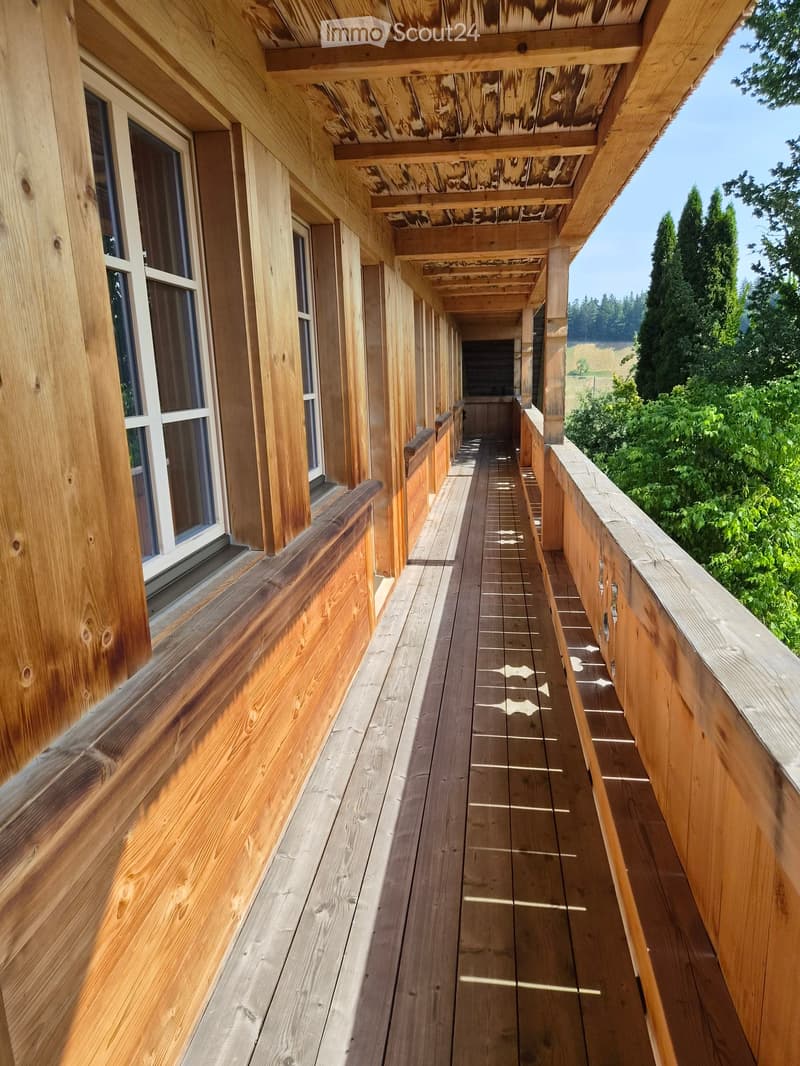 Wooden balcony with railings, wooden decking, and decorative patterns on the floor. The balcony is attached to a wooden building with large windows and overlooks a green landscape.