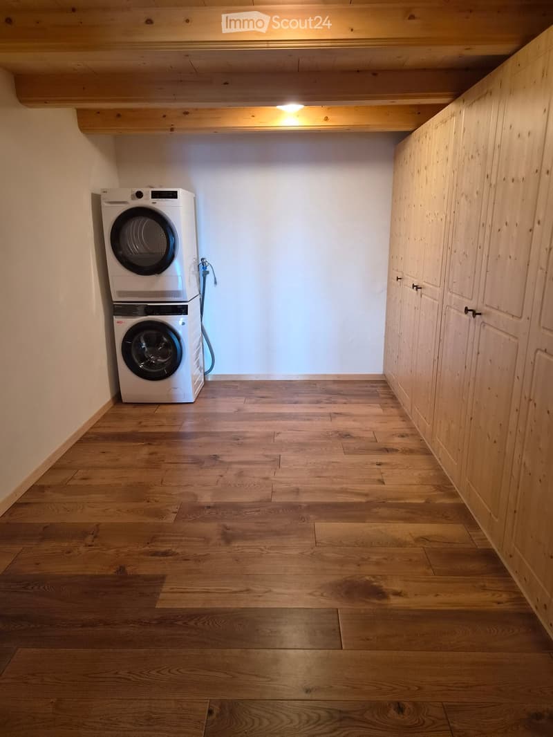 A small, well-lit room with wooden flooring and built-in wooden cabinets. Contains a stacked washing machine and dryer unit.