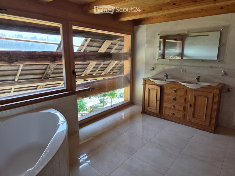 Bathroom with a large bathtub, double sink vanity, and a large mirror above the sink. The room features wooden walls and a large window offering a view of the outdoors.