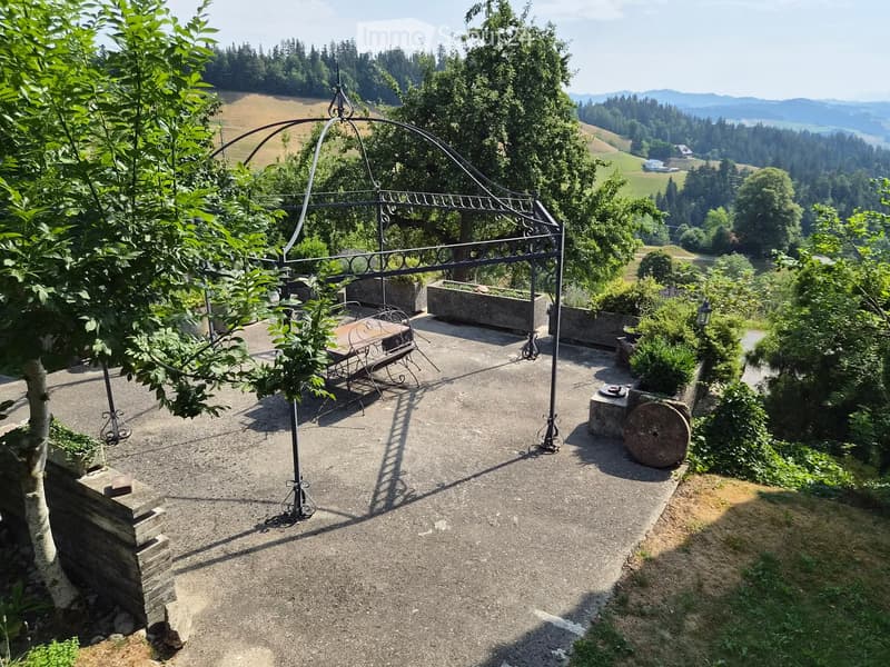Outdoor patio with a metal pergola, stone planters, a table and bench, surrounded by lush greenery and a scenic mountain view.