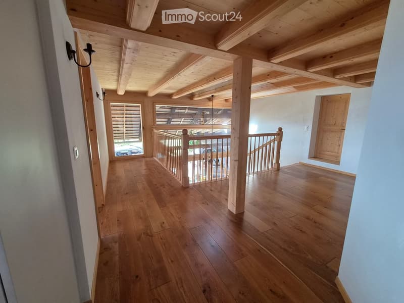 Spacious hallway with wooden flooring, exposed wooden beams on the ceiling, and a staircase leading to an upper level. The hallway features light blue walls, a wooden railing, and a large window with blinds.
